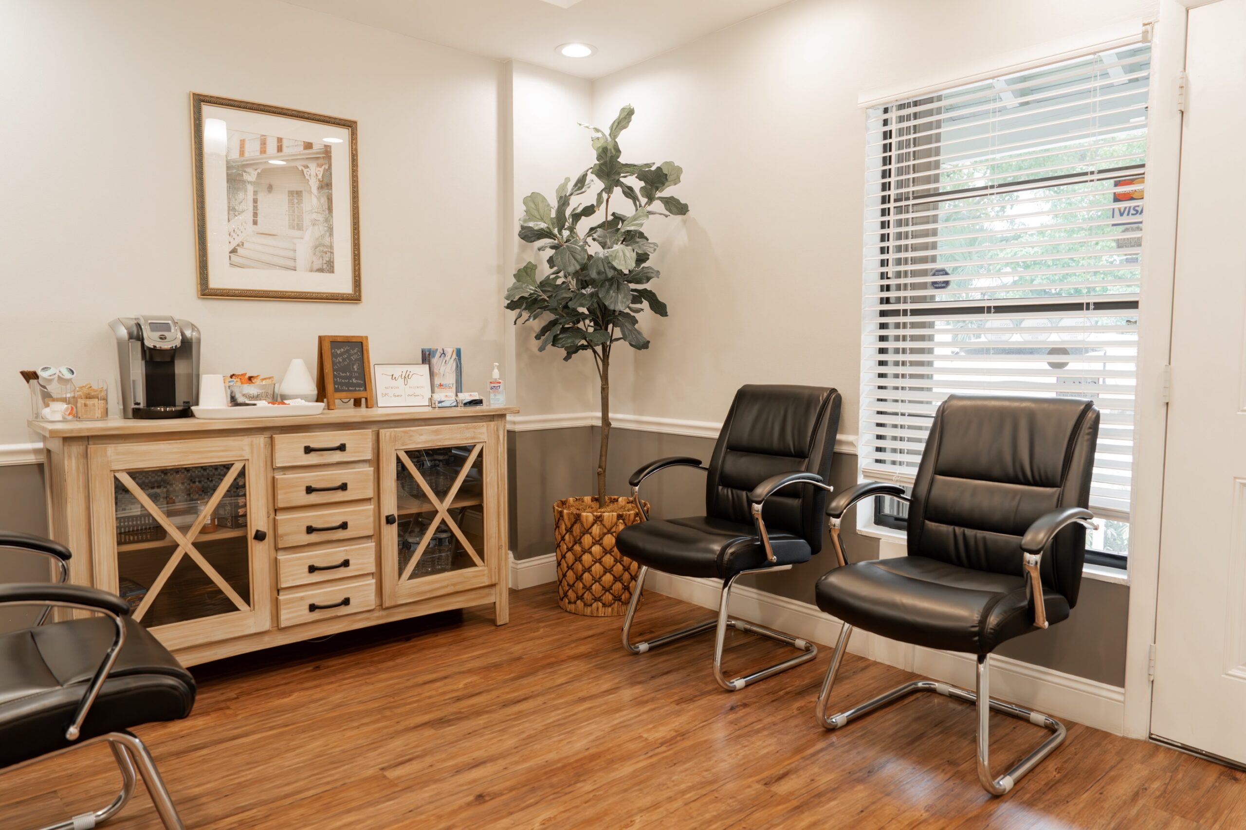 A clean and modern waiting room with two black leather chairs, a large wooden cabinet featuring glass panels and drawers, a coffee station with a Keurig, snacks, and hand sanitizer. A tall potted plant stands in the corner, while natural light streams through large windows with white blinds. The room has neutral-colored walls, wood flooring, and a framed photo on the wall, creating a welcoming and professional atmosphere.